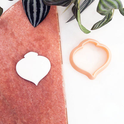A peach-colored silicone cookie cutter and a matching white cookie cutout displayed on a split white and pink background with decorative green leaves.