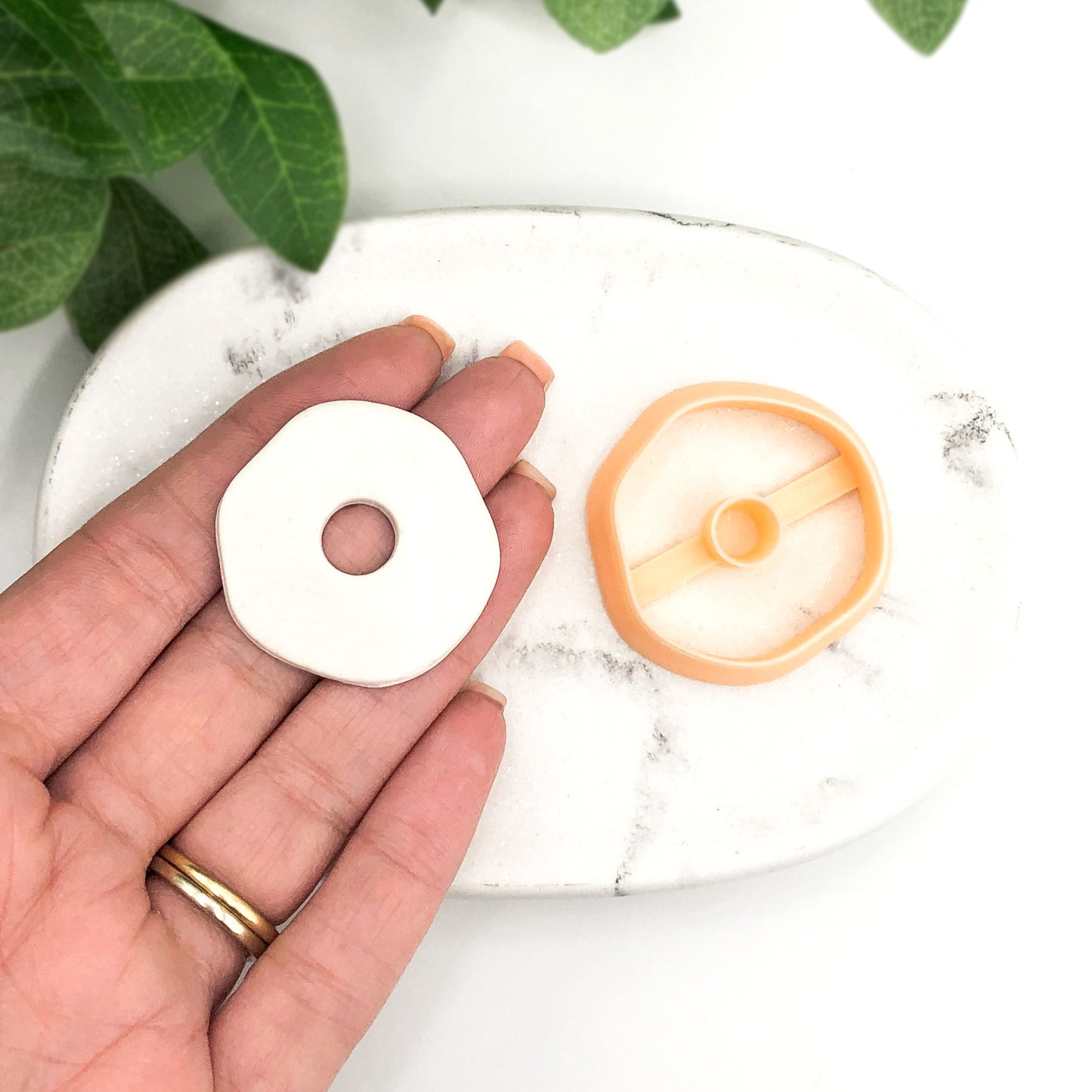 A hand holds a small white circular polymer clay cut-out piece with a centre hole next to an peach circular plastic 3d printed clay cutter on a white marble tray, with green leaves in the background.