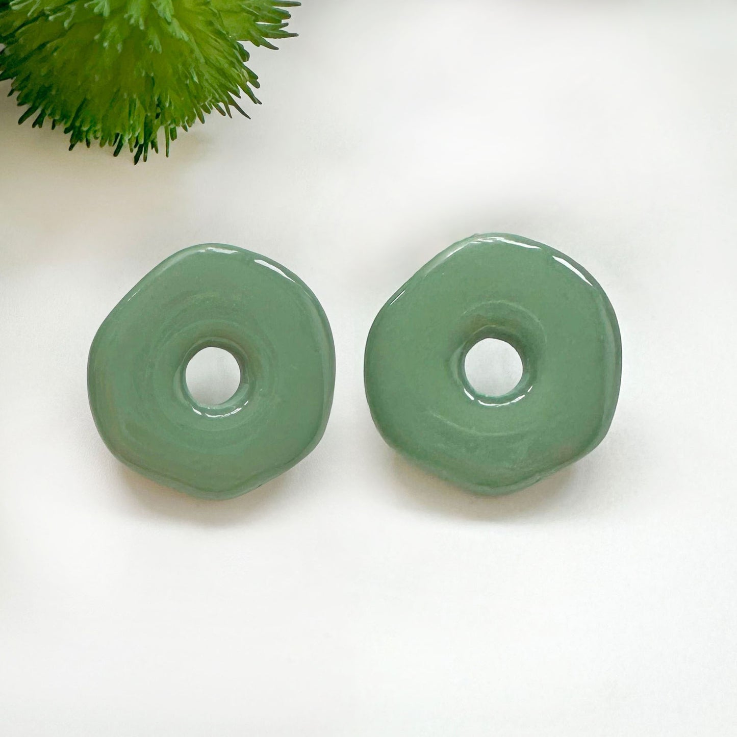 Two glossy sage green donut-shaped beads on a white background with a small green plant in the top-left corner.