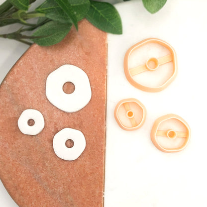 Three peach-colored plastic clay cookie cutters in different sizes are displayed on a white surface, with three of them shown pressing into white clay on a terracotta-colored stone slab and green leaves in the top left corner.