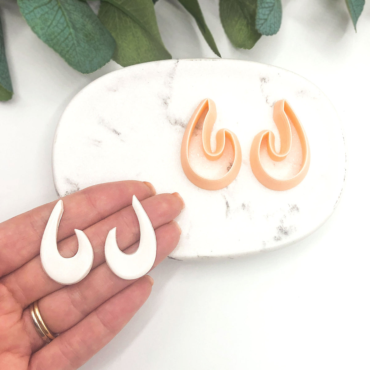 A pair of white organic droop-shaped clay cut outs held in a hand, with a matching pair of peach-colored clay cutters displayed on a white marble tray. Green leaves are visible in the background.