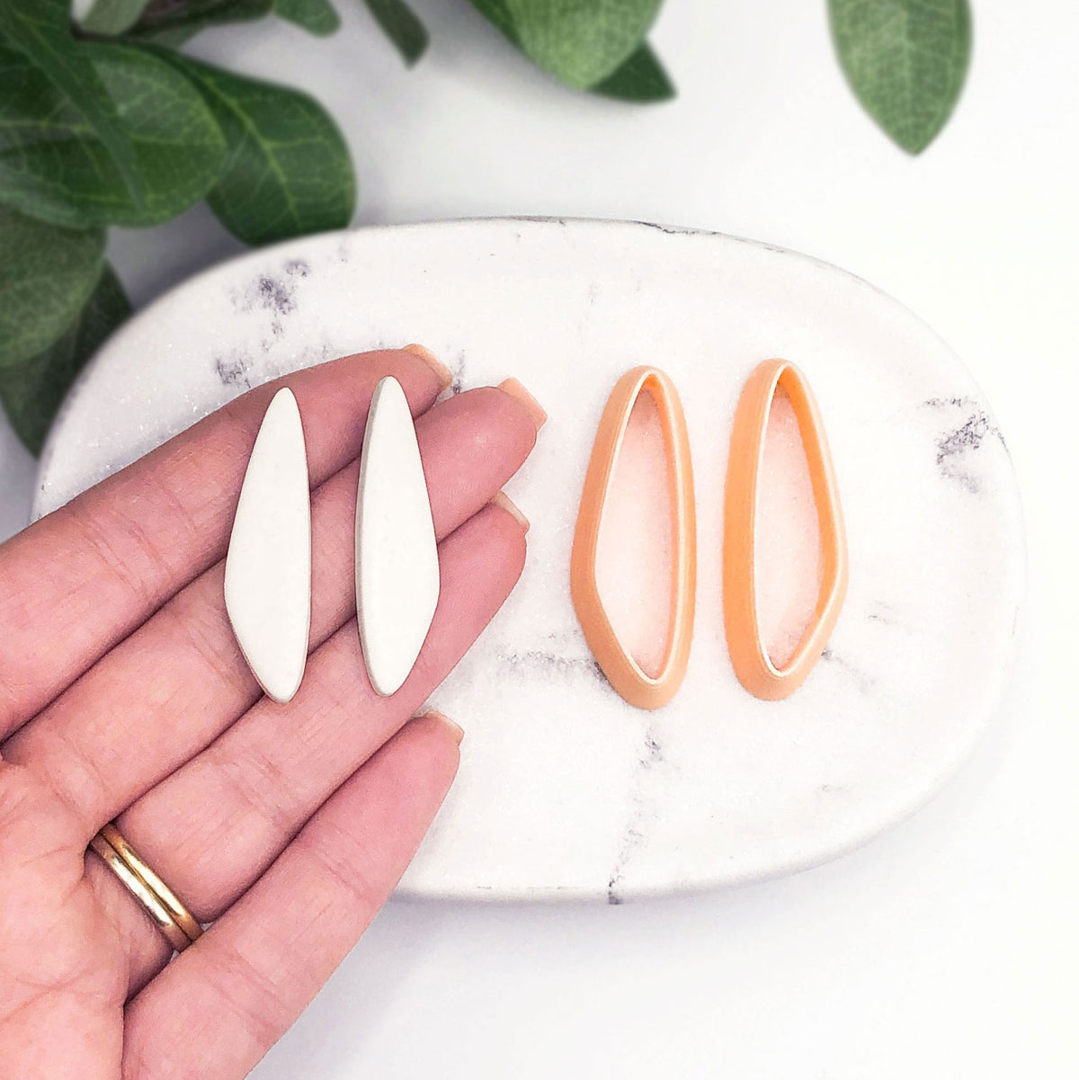 organic slim drop clay cutter, shown as a mirror set. Two clay cut outs on a hand then two mirrored peach clay cutters on a marble tray with green leaves in the background.