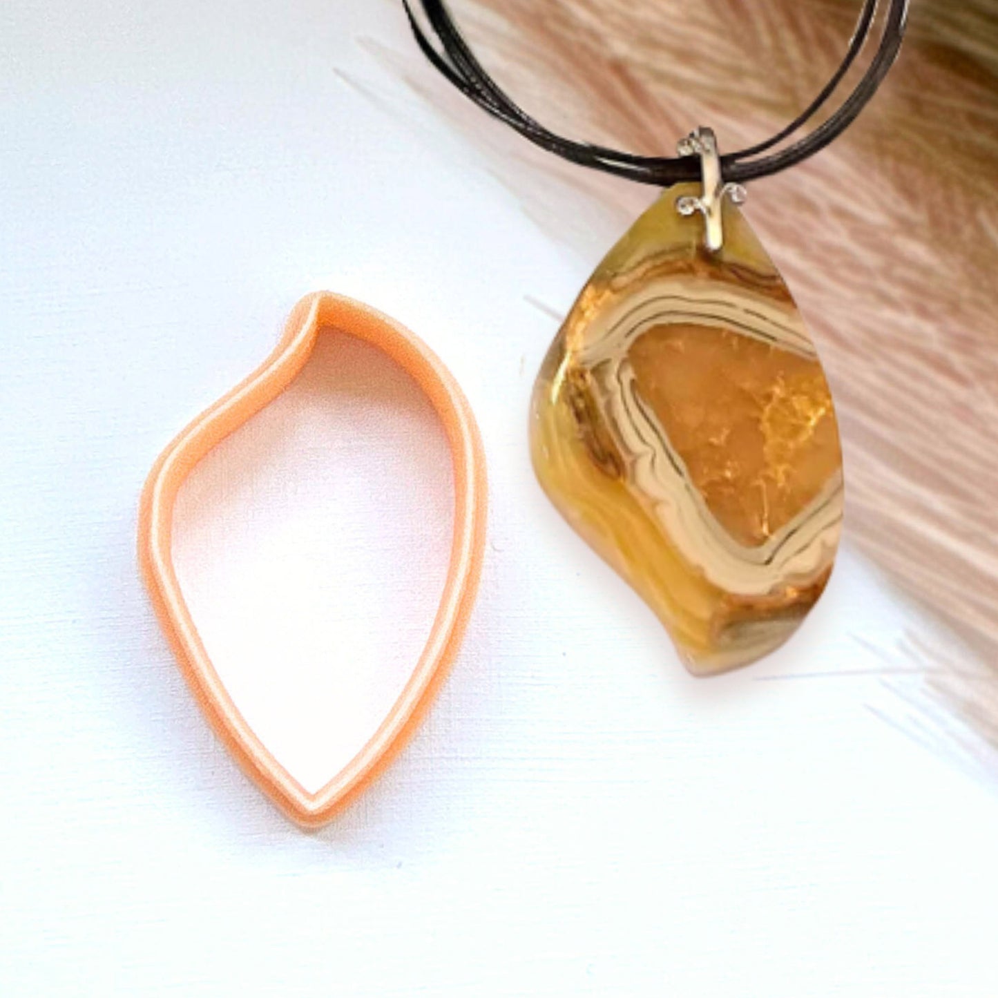 A teardrop-shaped polymer clay amber agate geode pendant necklace on a black cord, shown next to its peach-colored clay cutter on a white background.
