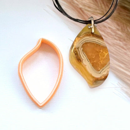 A teardrop-shaped polymer clay amber agate geode pendant necklace on a black cord, shown next to its peach-colored clay cutter on a white background.