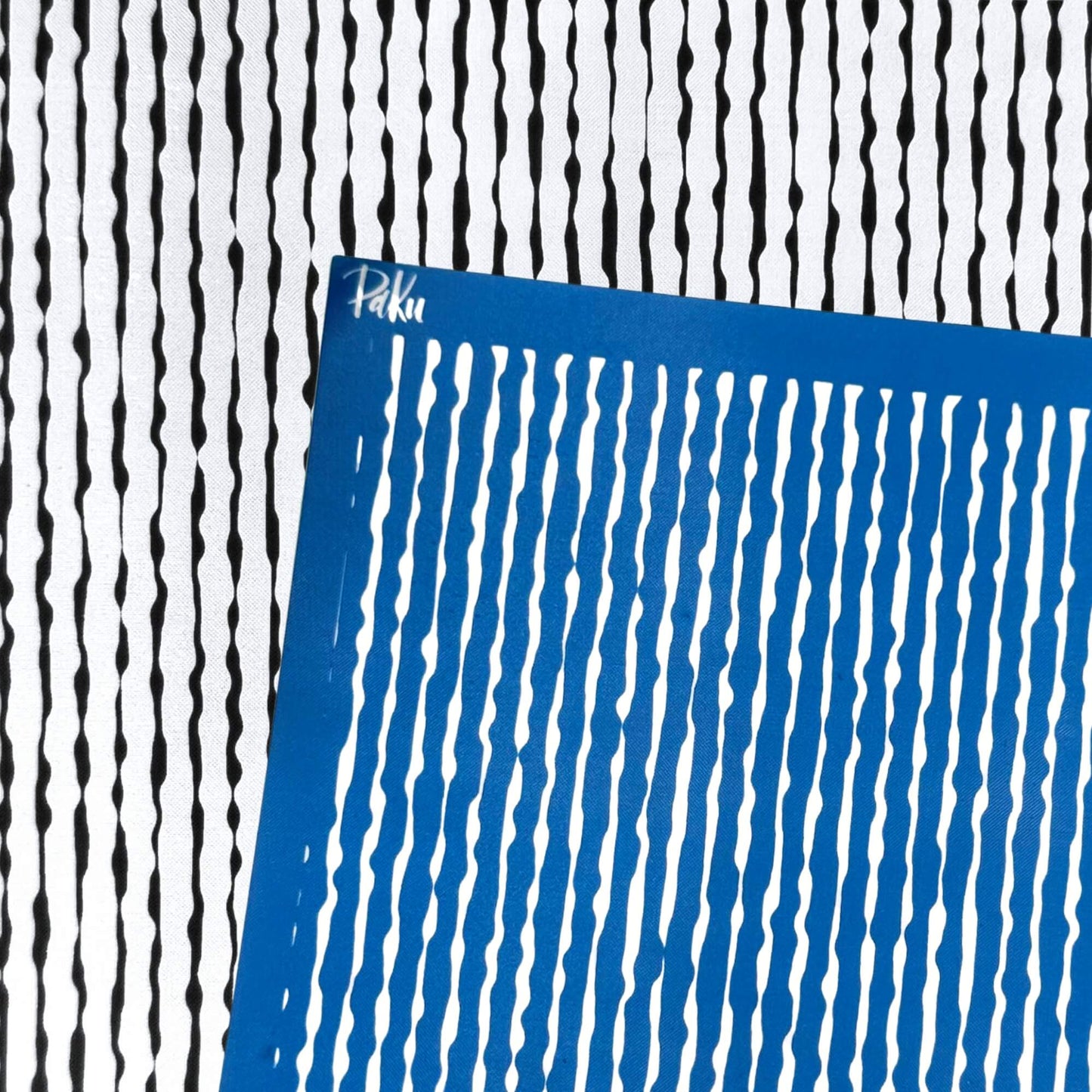 A blue square silkscreen stencil with vertical wavy organic lines, placed diagonally over a black and white striped patterned background. The brand name 'Paku' is visible in white script on the top-left corner of the blue sheet.