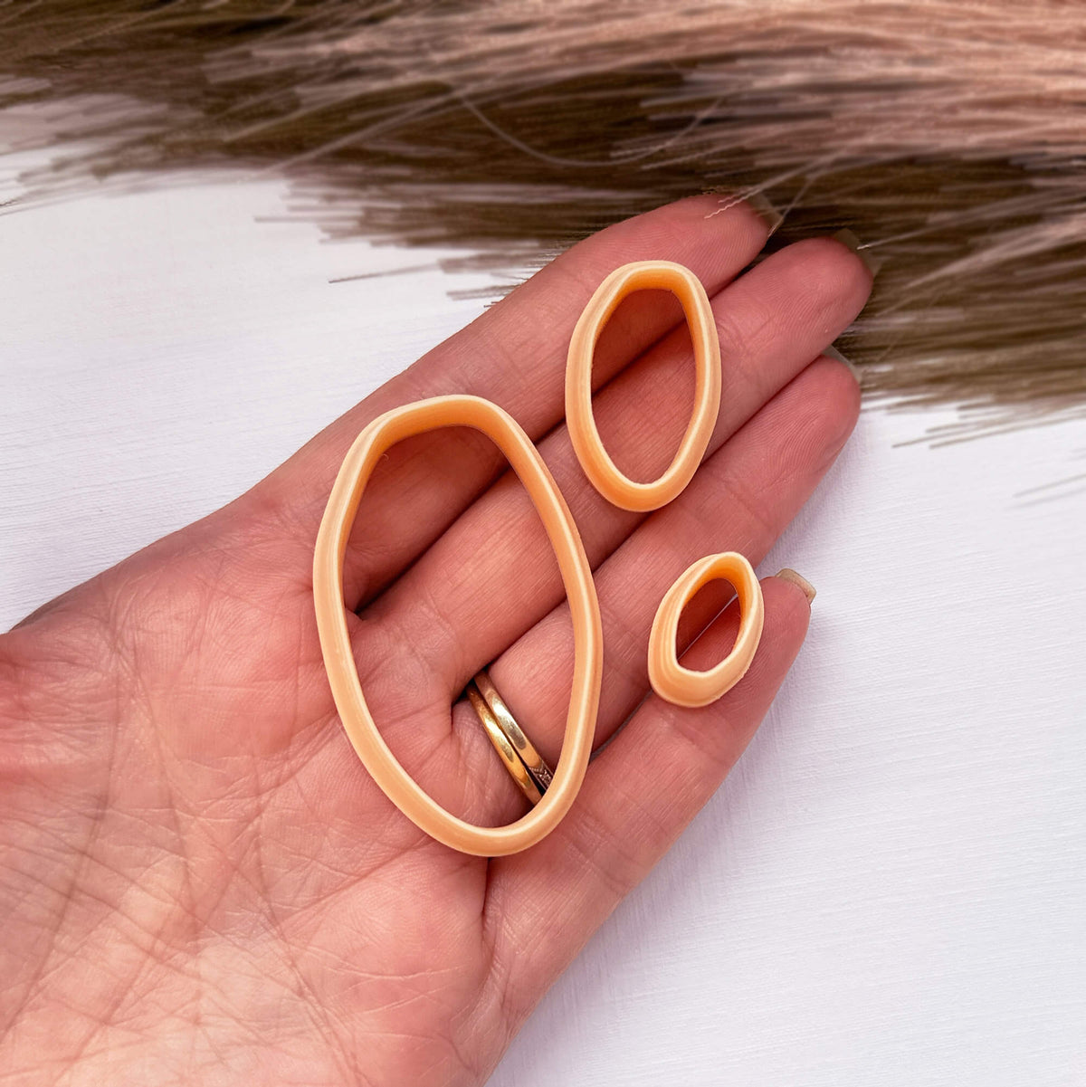 Three peach-colored clay cutters of different sizes held in a person's hand, with a gold ring on the finger, against a white background 