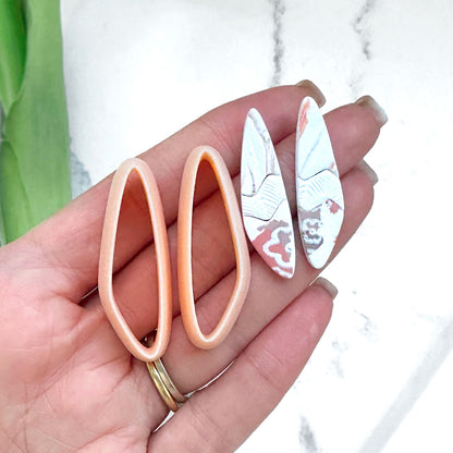 organic slim drop clay cutter, shown as a mirror set. Two clay cut outs on a hand and two mirrored peach clay cutters with green leaves in the background.