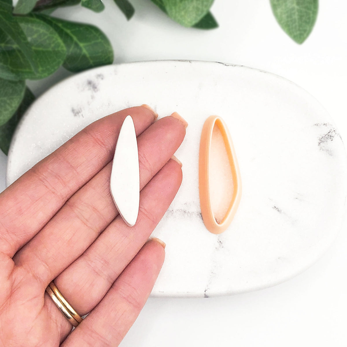 organic slim drop clay cutter. A clay cut out is on a hand then the peach clay cutters on a marble tray with green leaves in the background.