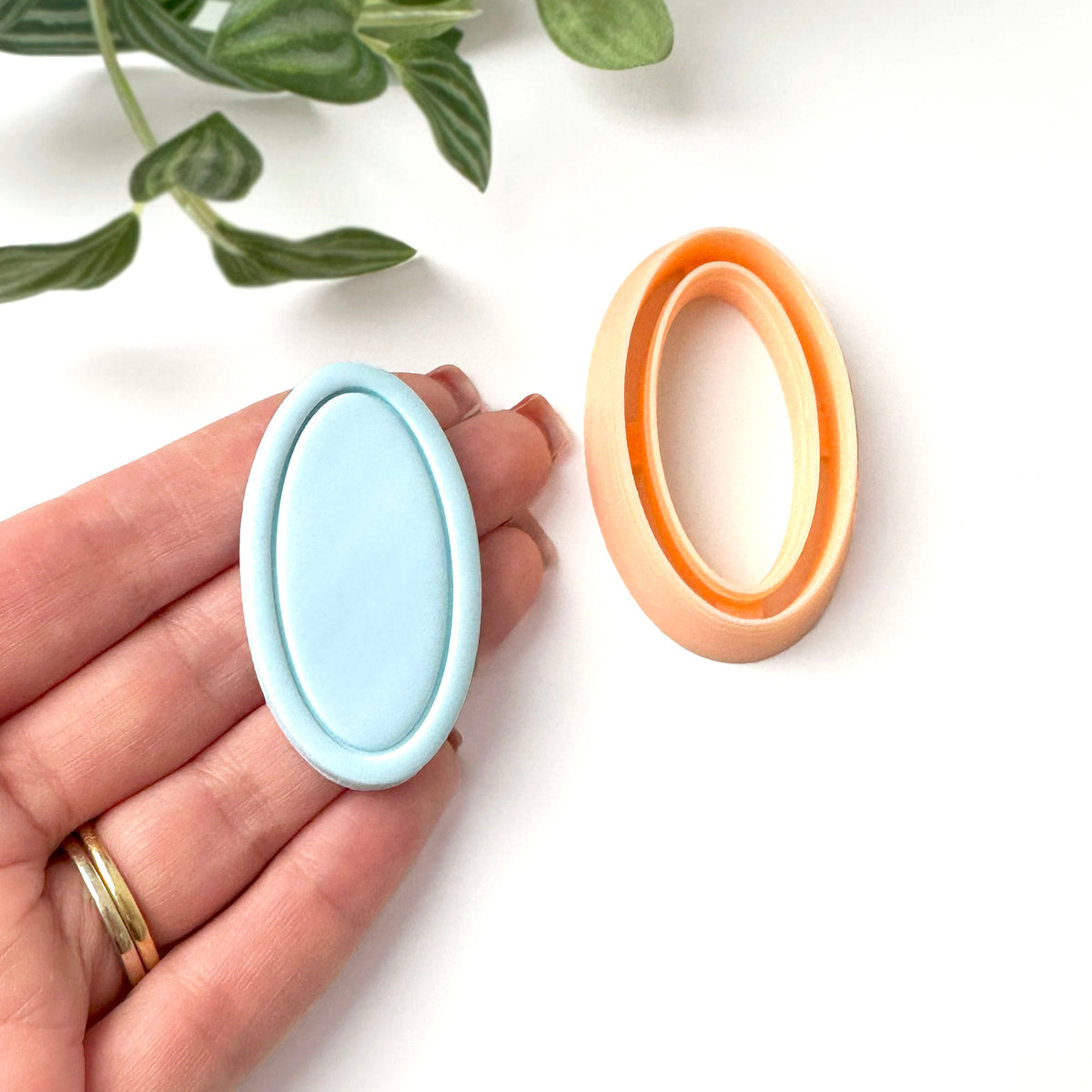 A light blue oval-shaped polymer clay cut out with a framed edge. The mould is held in a hand. A  peach-coloured oval-shaped clay cutter is also shown.