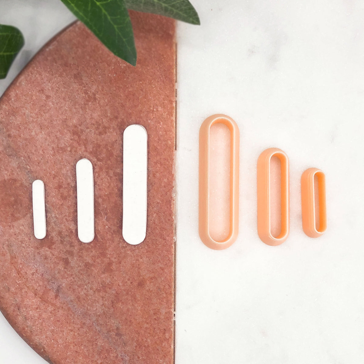 Four oval-shaped cookie cutters in peach and orange tones arranged on a white marble surface next to a copper-colored stone slab with matching white cutouts, with green leaves in the top corner.