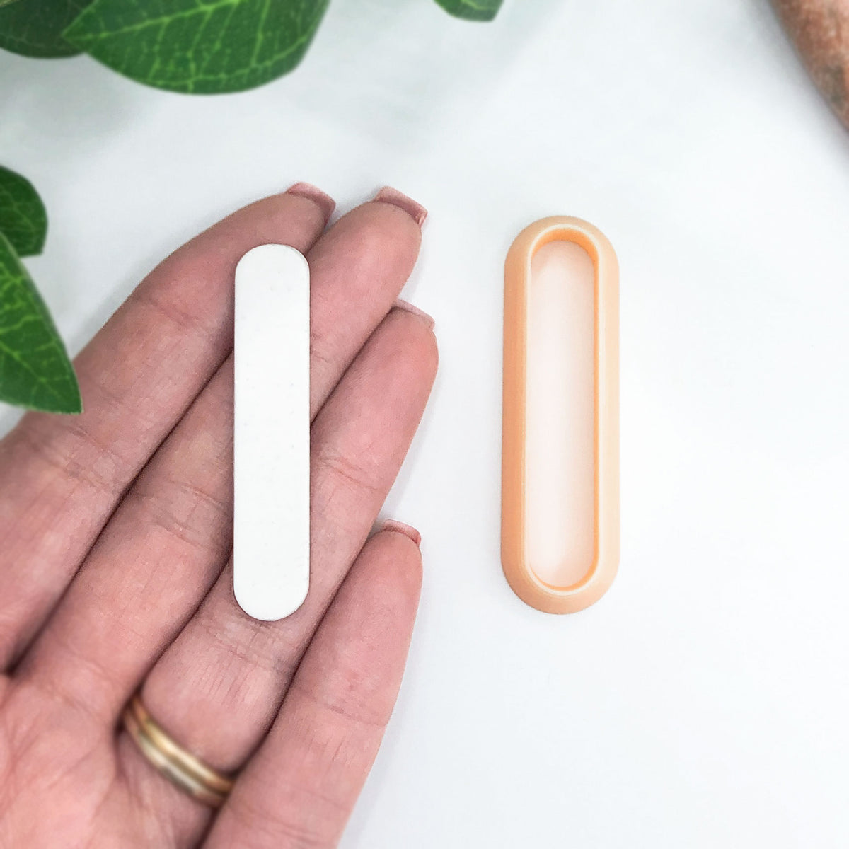 A hand holds a small white oval-shaped clay cut out next to a peach-colored clay cutter, shown on a clean white surface with green leaves in the background.