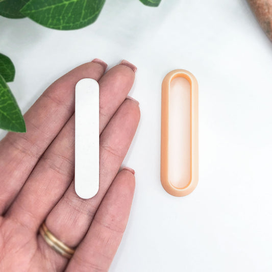 A hand holds a small white oval-shaped clay cut out next to a peach-colored clay cutter, shown on a clean white surface with green leaves in the background.