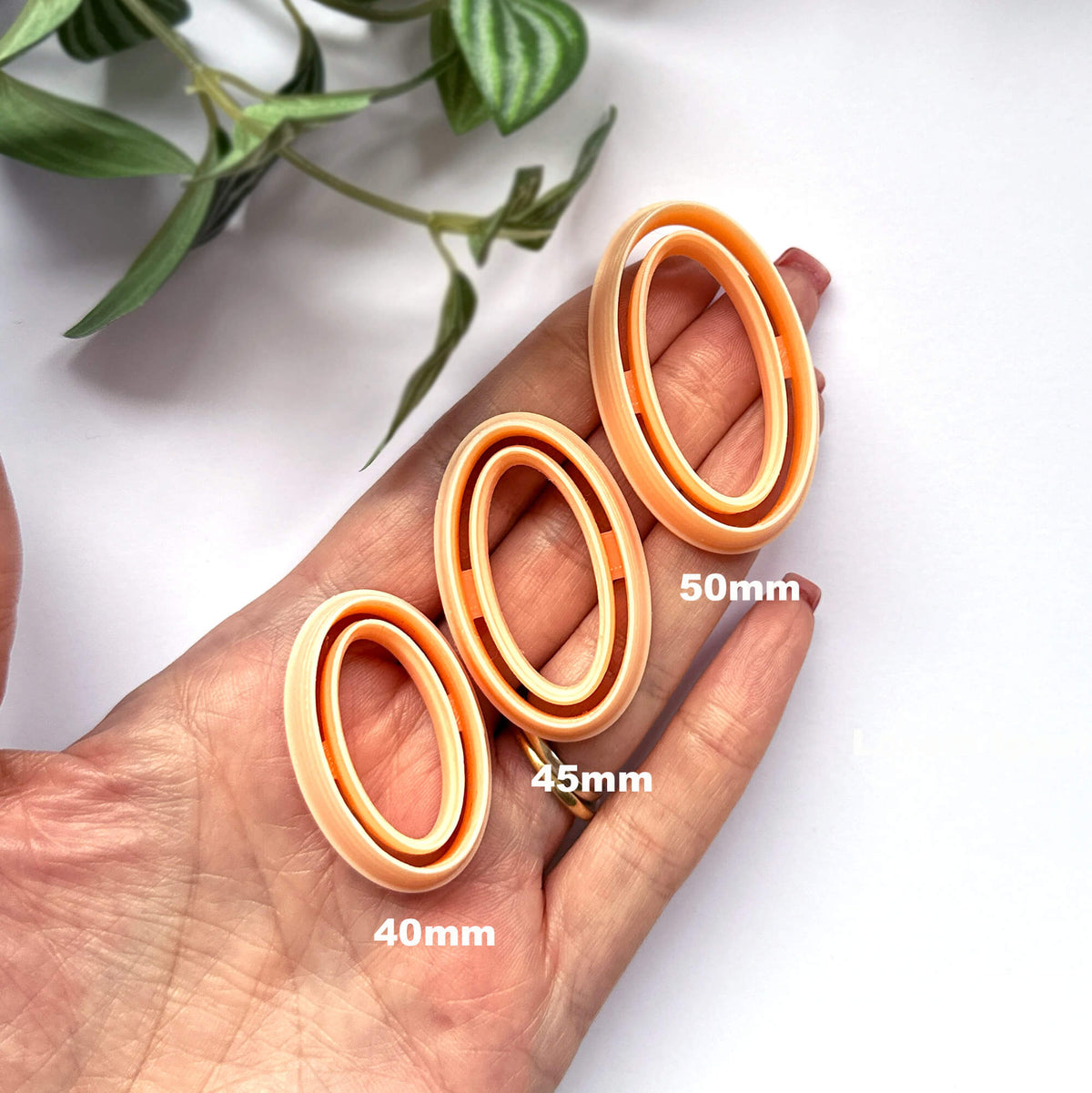 Three peach-colored oval frame clay cutters displayed on a person's hand, labeled with sizes 40mm, 45mm, and 50mm, with green leaves in the background.