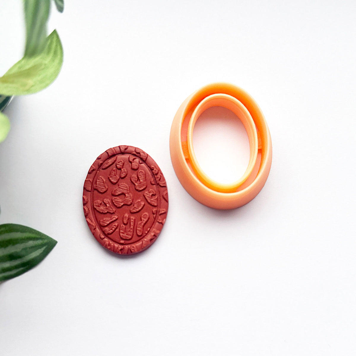 An oval orange frame clay cutter and a matching red clay cut out impression on a clean white background with decorative green leaves in the corner.
