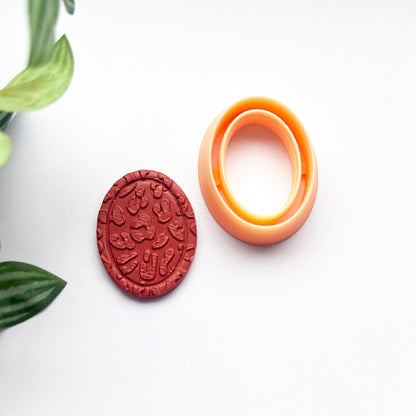 An oval orange frame clay cutter and a matching red clay cut out impression on a clean white background with decorative green leaves in the corner.