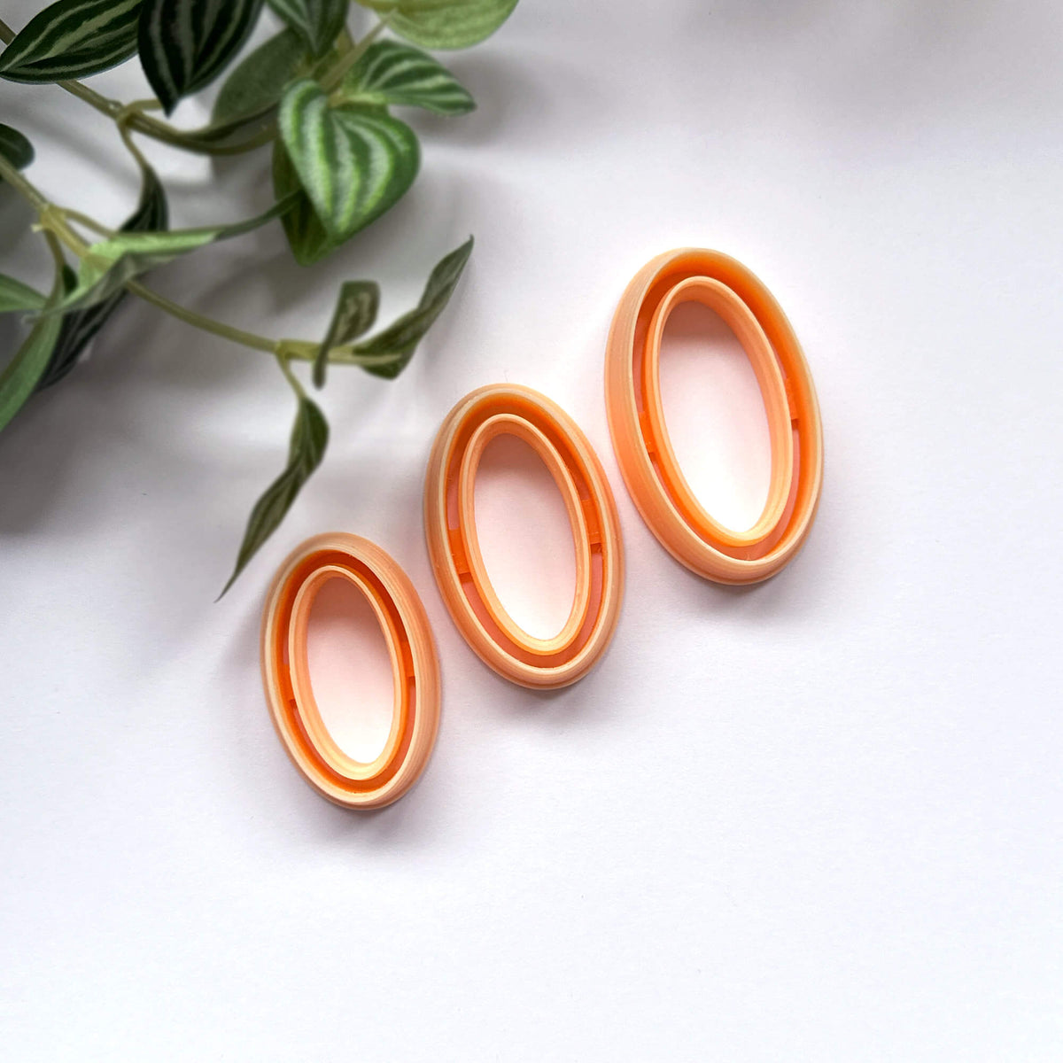 Three oval-shaped orange clay cutters arranged diagonally on a white surface next to a green plant.