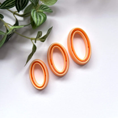 Three oval-shaped orange clay cutters arranged diagonally on a white surface next to a green plant.
