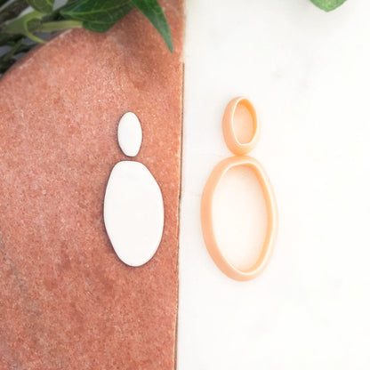A peach-colored plastic cookie clay cutter shaped organic oval is shown next to two white clay cutouts on a split white and terracotta background.