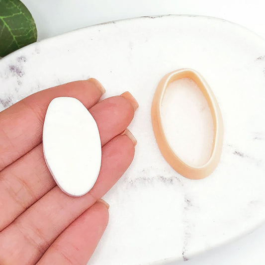 A hand holds a small white oval organic polymer clay cut out next to a peach oval clay cutter on a white marble surface.