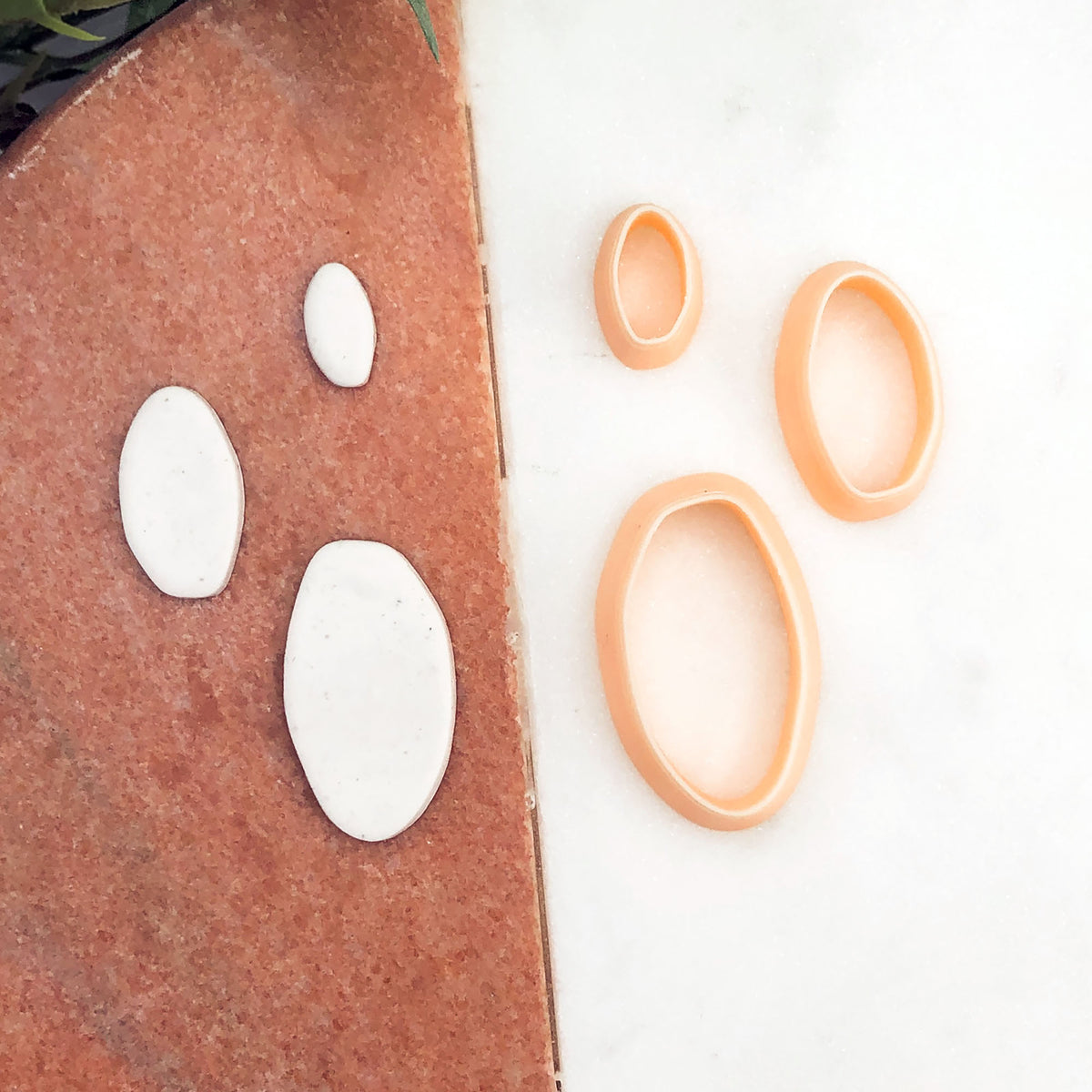 three peach-colored oval cookie cutters are displayed on a white marble surface next to four matching white clay cutouts on a terracotta surface.