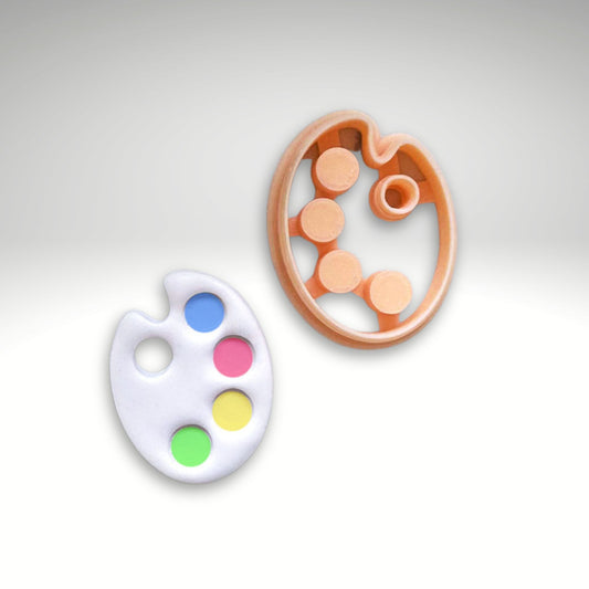 3d printed paint palette-shaped clay cutter on a light gray gradient background: polymer clay cut out white piece with blue, pink, yellow, and green color wells