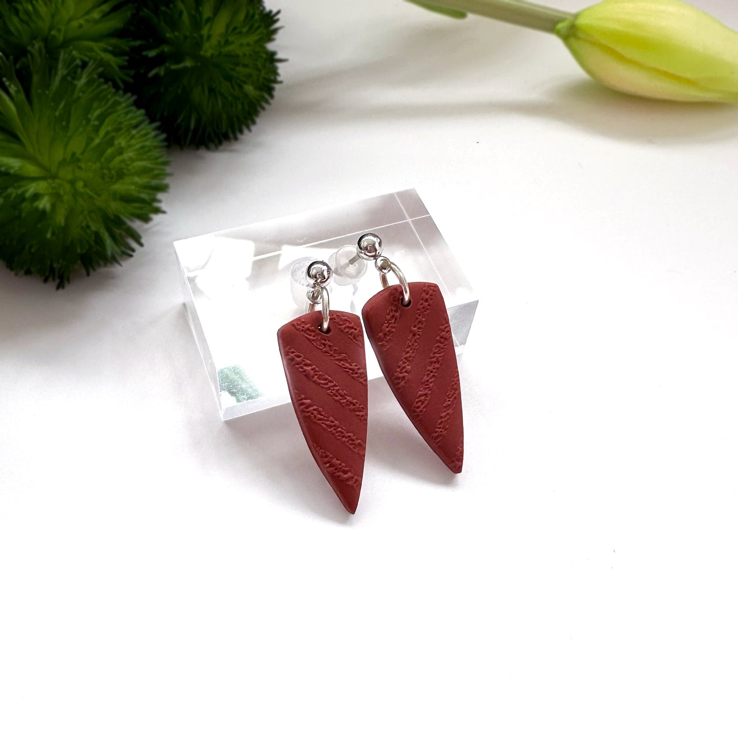 Red polymer clay earrings on a clear stand with green foliage and a yellow flower in the background