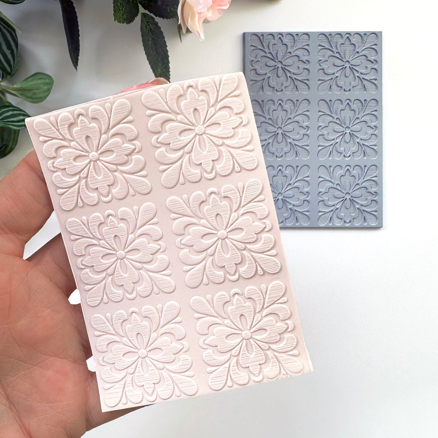 A hand holds a pale pink rectangular  sheet of polymer clay with embossed floral motifs of portuguese spanish tiles with a gray rubber texture sheet placed behind it on a white surface. Decorative leaves and a flower are partially visible at the top.