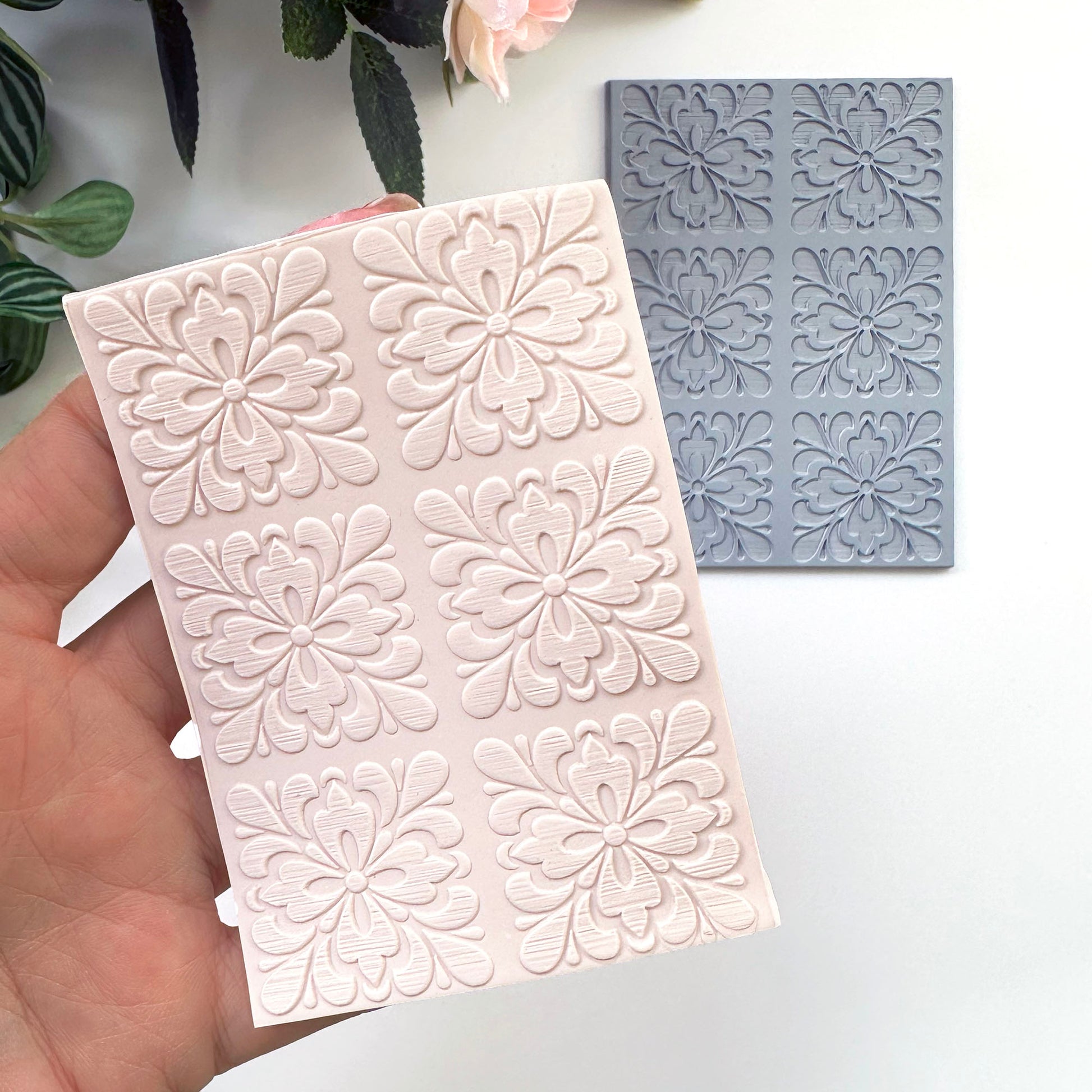 A hand holds a pale pink rectangular  sheet of polymer clay with embossed floral motifs of portuguese spanish tiles with a gray rubber texture sheet placed behind it on a white surface. Decorative leaves and a flower are partially visible at the top.