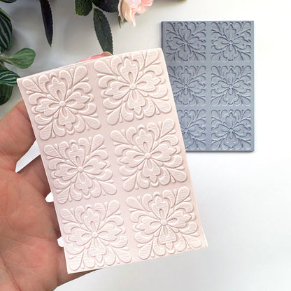 A hand holds a pale pink rectangular  sheet of polymer clay with embossed floral motifs of portuguese spanish tiles with a gray rubber texture sheet placed behind it on a white surface. Decorative leaves and a flower are partially visible at the top.