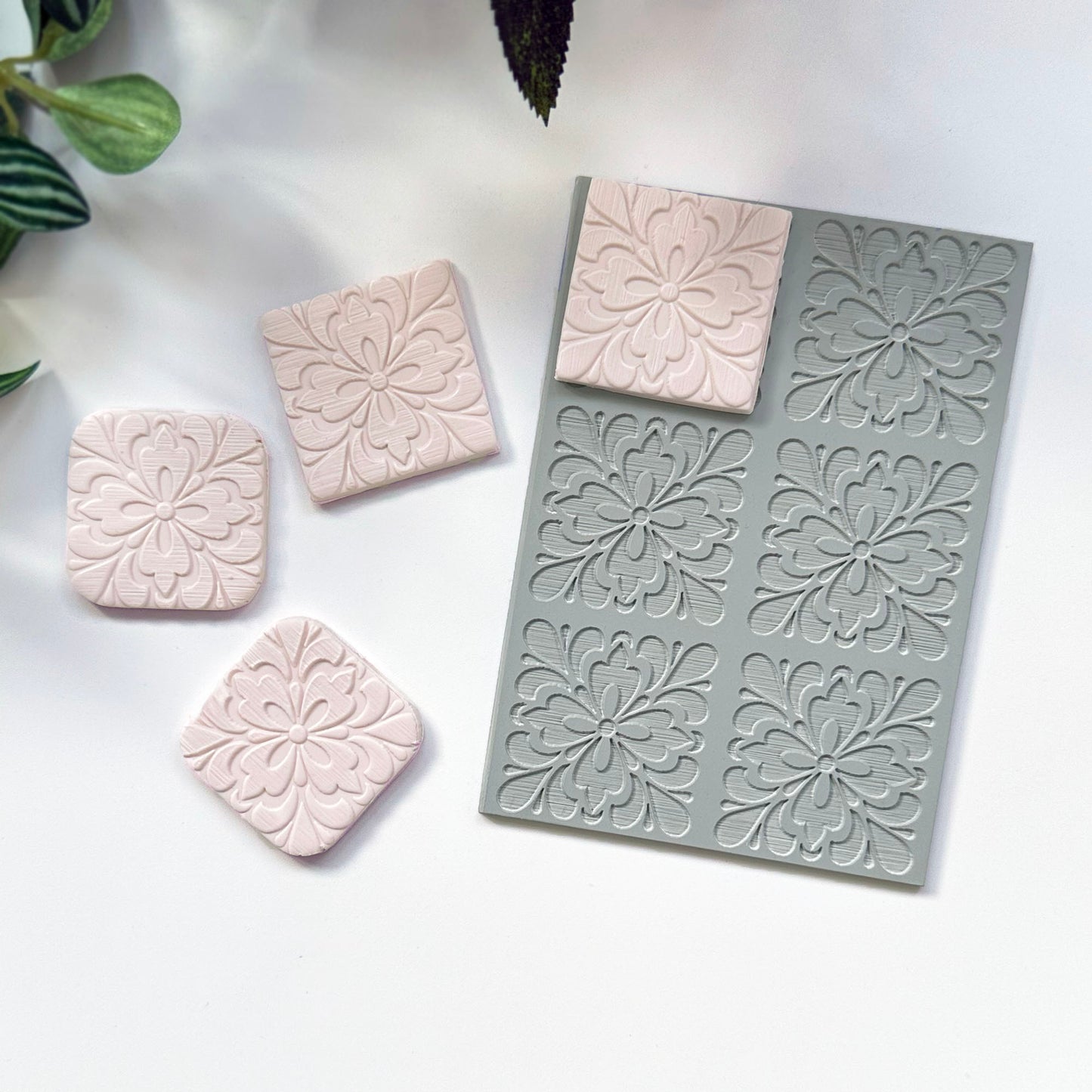 Pale pink squares of polymer clay with embossed floral motifs of portuguese spanish tiles with a gray rubber texture sheet placed behind it on a white surface. Decorative leaves and a flower are partially visible at the top.