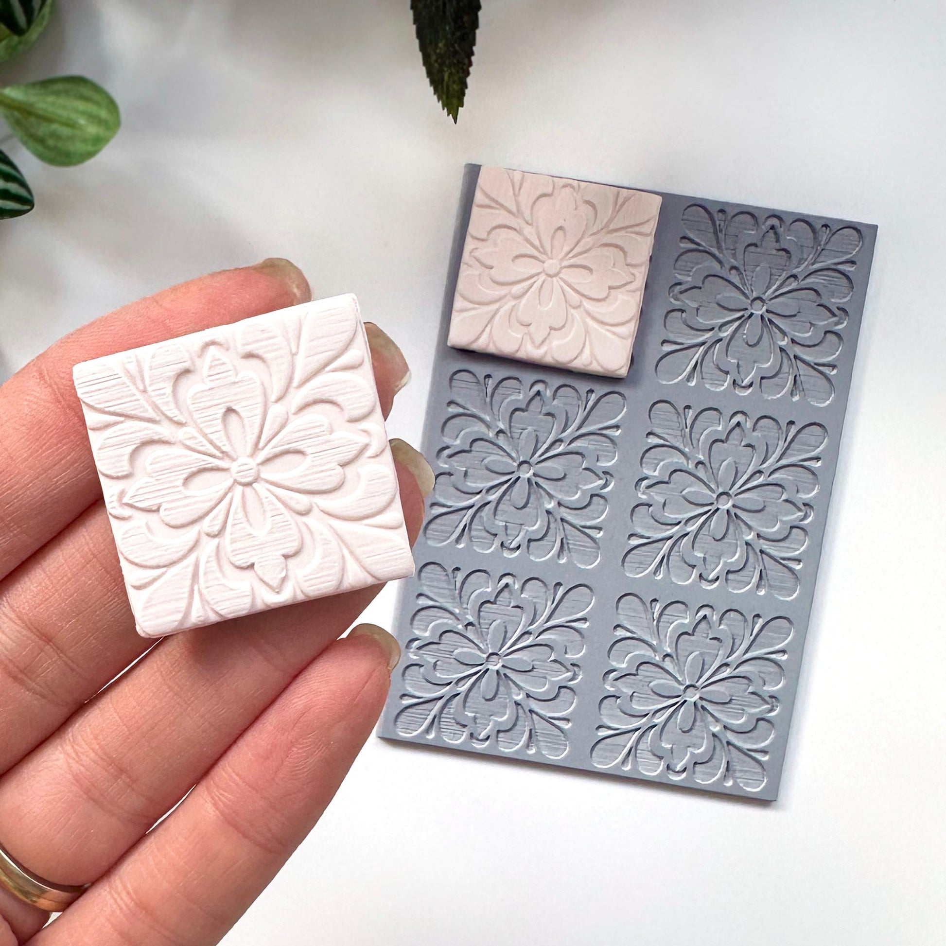 A hand holds a small square white polymer clay piece with an embossed floral pattern next to a gray silicone stamp showing the same floral impression, on a clean white background.