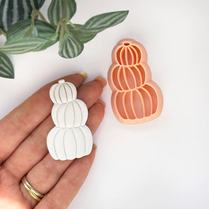 A hand holds a small white pumpkin-shaped clay cutout next to a peach-colored pumpkin-shaped cookie clay cutter on a clean white background, with green leaves in the top left corner.