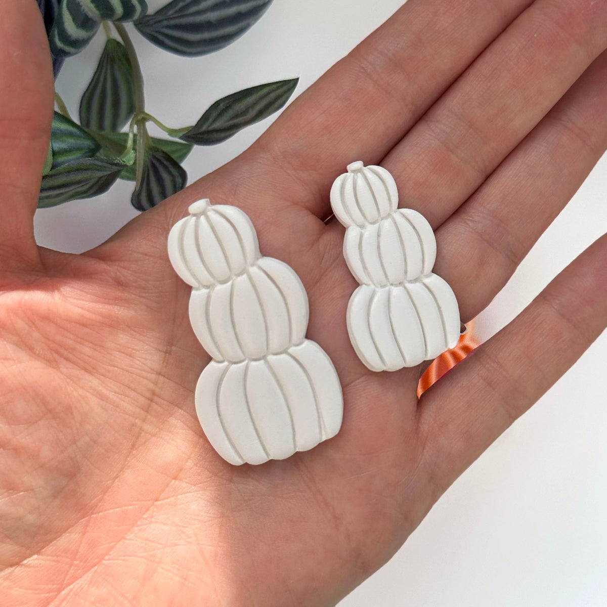 A pair of white stacked pumpkin-shaped clay cut outs resting on an open hand, with a green leafy plant in the background.