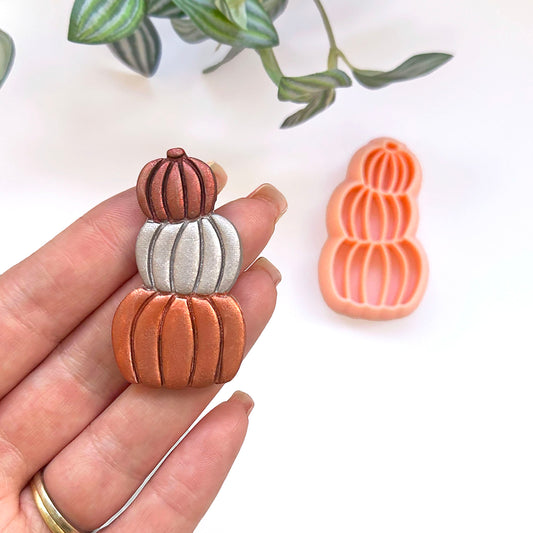 A hand holds a small pumpkin-shaped accessory with a stacked pumpkin design in earthy tones, while a matching orange clay cutter is shown on a white surface with blurred green leaves in the background.