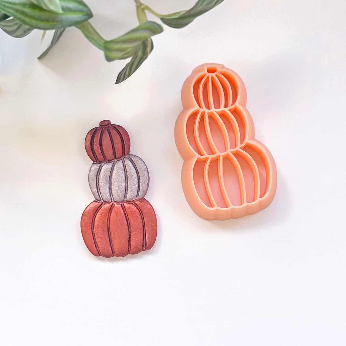 A 3d printed pumpkin-shaped cookie cutter in peach-orange next to a matching cutout in red, orange, and purple, displayed on a clean white surface with a decorative green plant leaf in the top-left corner.
