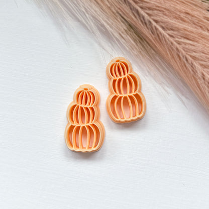 Two mirrored small orange pumpkin-shaped cookie clay cutters on a white textured surface with dried beige grass in the background.