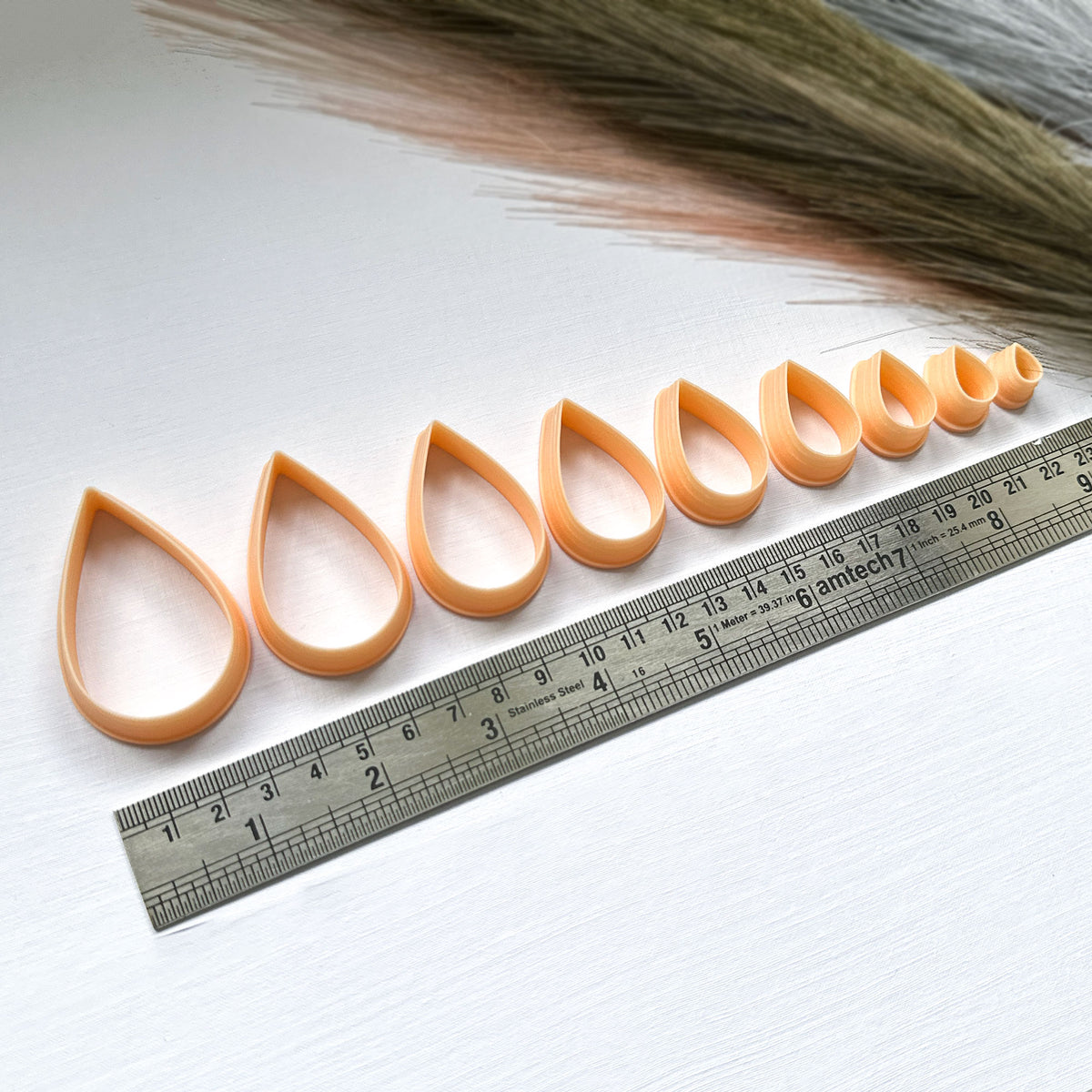 A set of nine peach-coloured plastic clay cutters in various sizes, ranging from 10mm to 50mm. The cutters are shaped like raindrops and have a ruler under them for size comparison.