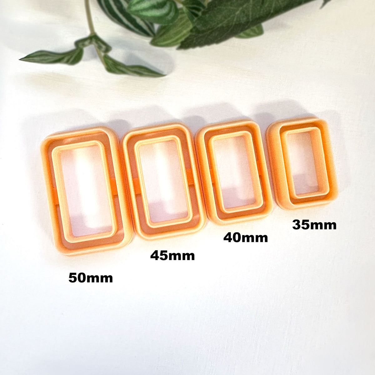 Four rectangular cookie clay cutters in orange and white, shown in sizes 50mm, 45mm, 40mm, and 35mm, arranged in descending size order on a white background with green leaves in the top corner.