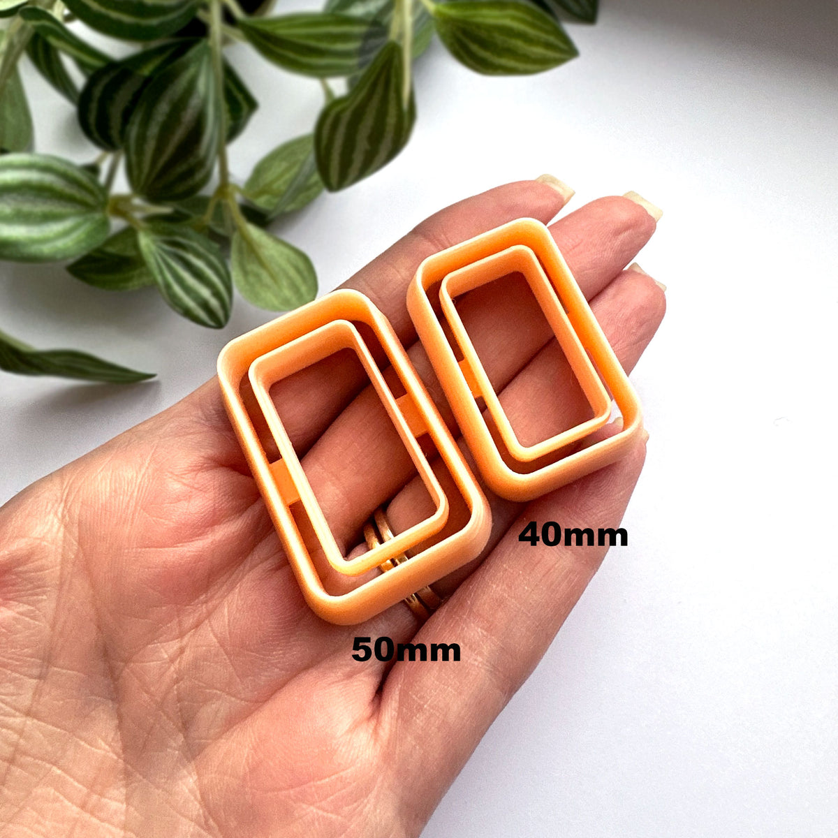 Two small peach clay cutter rectangular plastic frames held in a person's hand, with green leaves in the upper left and a white background.