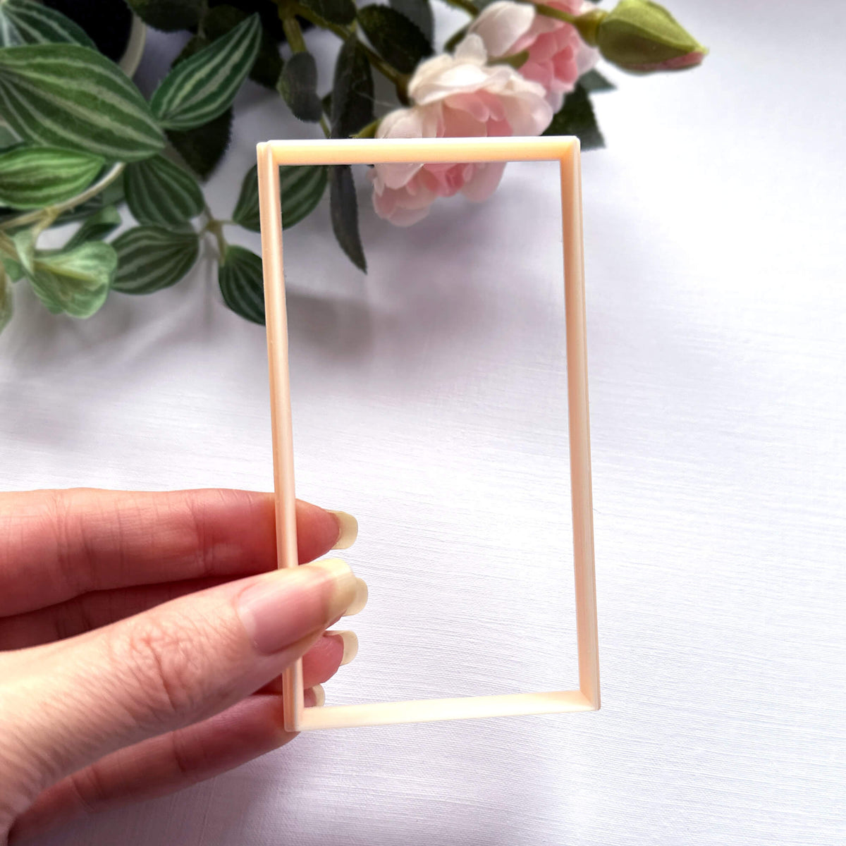 A hand holding a rectangular plastic clay cutter in a light peach colour, set against a white background with green leaves and pink flowers in the upper corner.