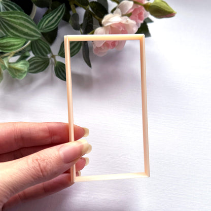 A hand holding a rectangular plastic clay cutter in a light peach colour, set against a white background with green leaves and pink flowers in the upper corner.