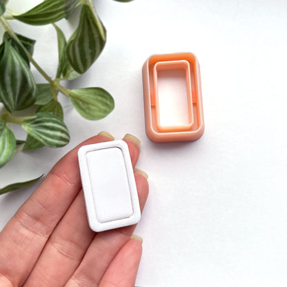 Rounded rectangle clay cutter sits on a white background while a hand holds a white polymer cut out made using the cutter.