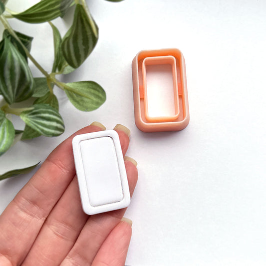 Rounded rectangle clay cutter sits on a white background while a hand holds a white polymer cut out made using the cutter.