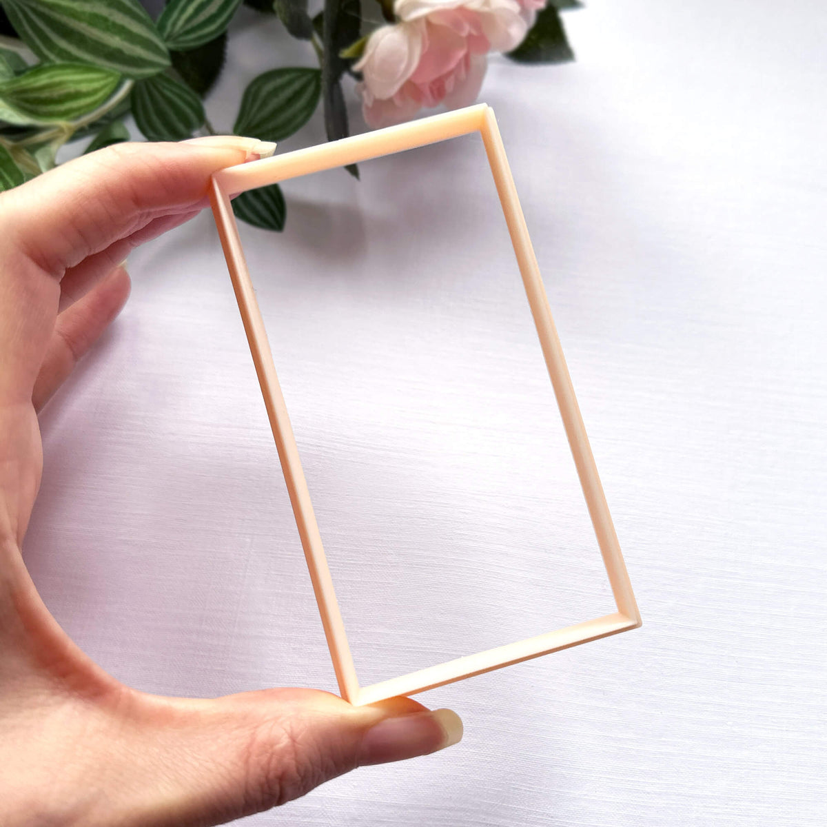 A hand holding a clear rectangle clay cutter in a thin peach colour, set against a white textured background with soft pink flowers in the upper corner.