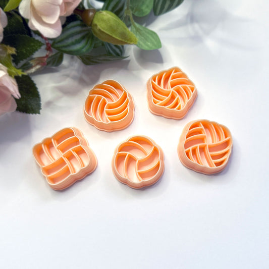Five peach-colored plastic cookie clay cutters with different knot geometric and floral-inspired patterns, arranged on a white surface with decorative pink flowers in the top left corner.