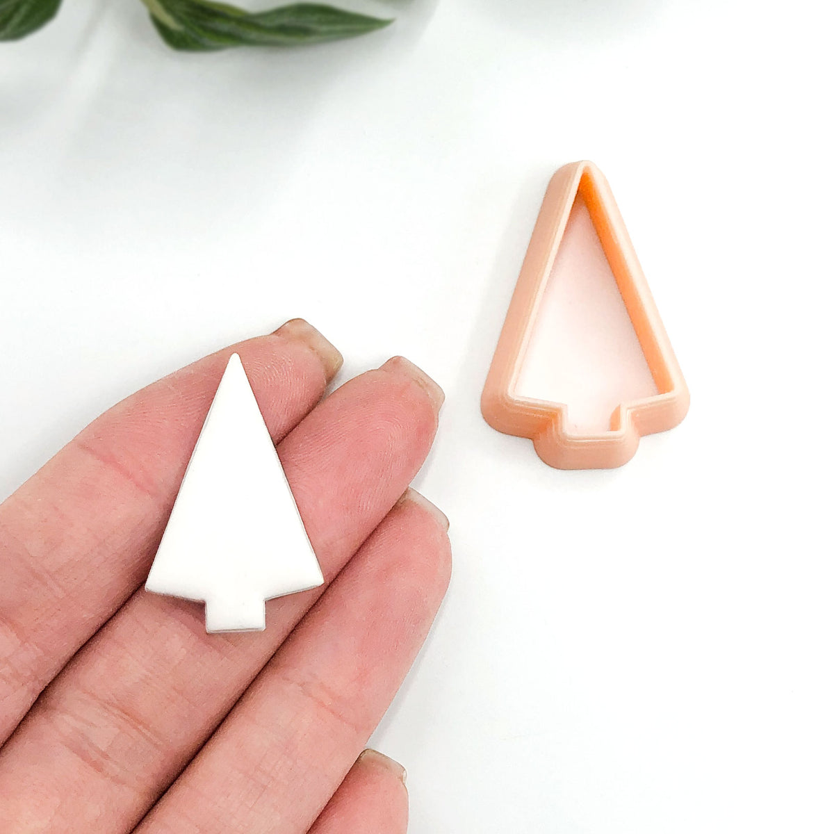 A hand holds a small white tree-shaped item while a peach-colored tree-shaped cookie cutter sits on a white surface nearby.