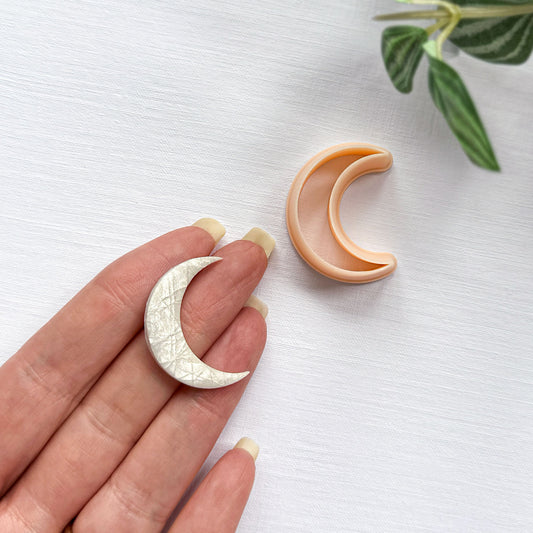 A hand holds a small crescent moon-shaped clay charm. Next to a peach-colored crescent moon clay cutter on a white textured background with green leaves in the corner.