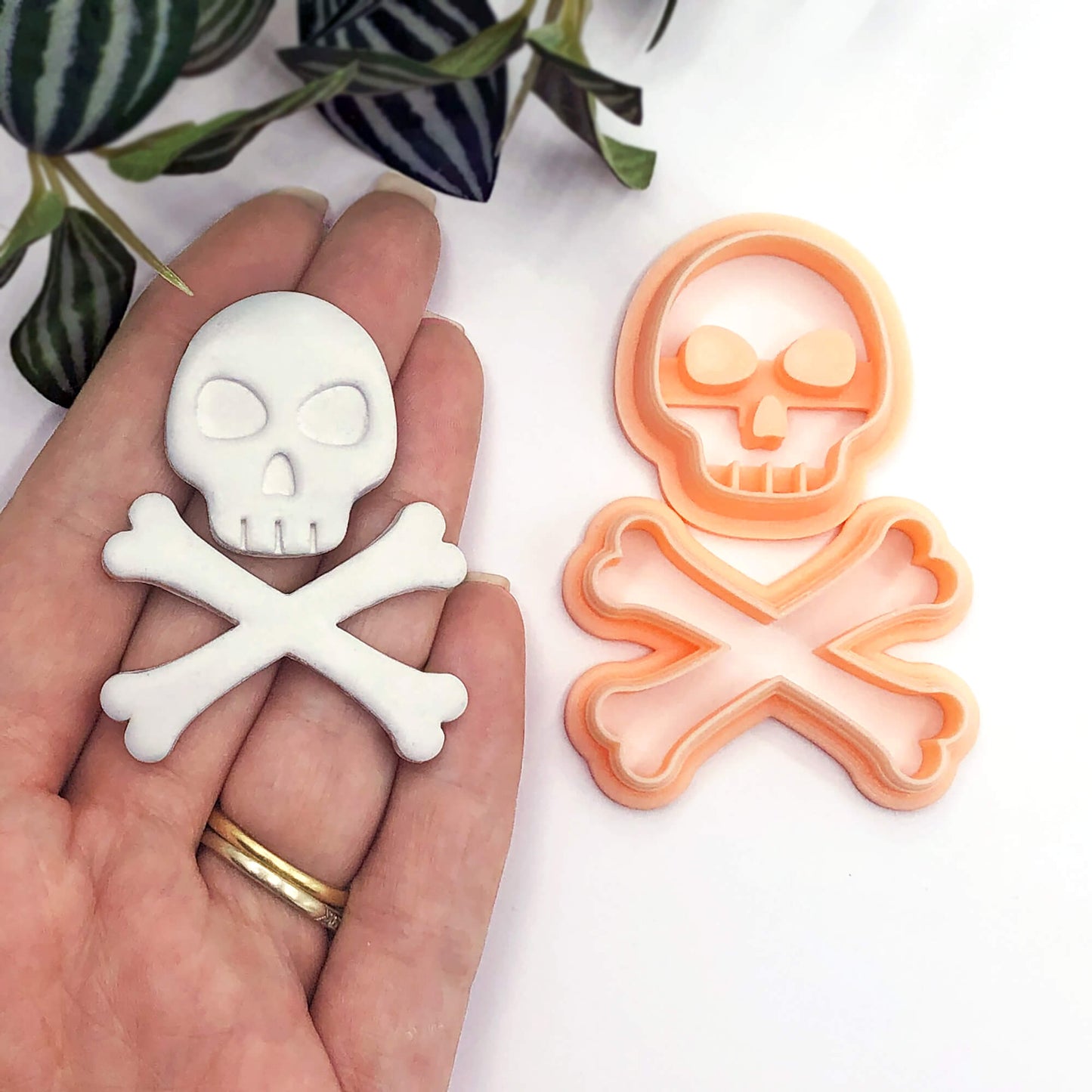A hand holds a white skull-and-crossbones-shaped polymer clay item, with an orange matching cookie cutter shown beside it on a white surface. Green leaves are visible in the upper left corner.