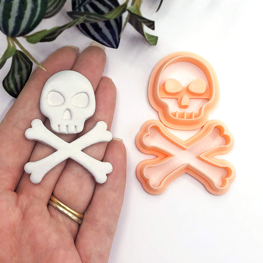 A hand holds a white skull-and-crossbones-shaped polymer clay item, with an orange matching cookie cutter shown beside it on a white surface. Green leaves are visible in the upper left corner.