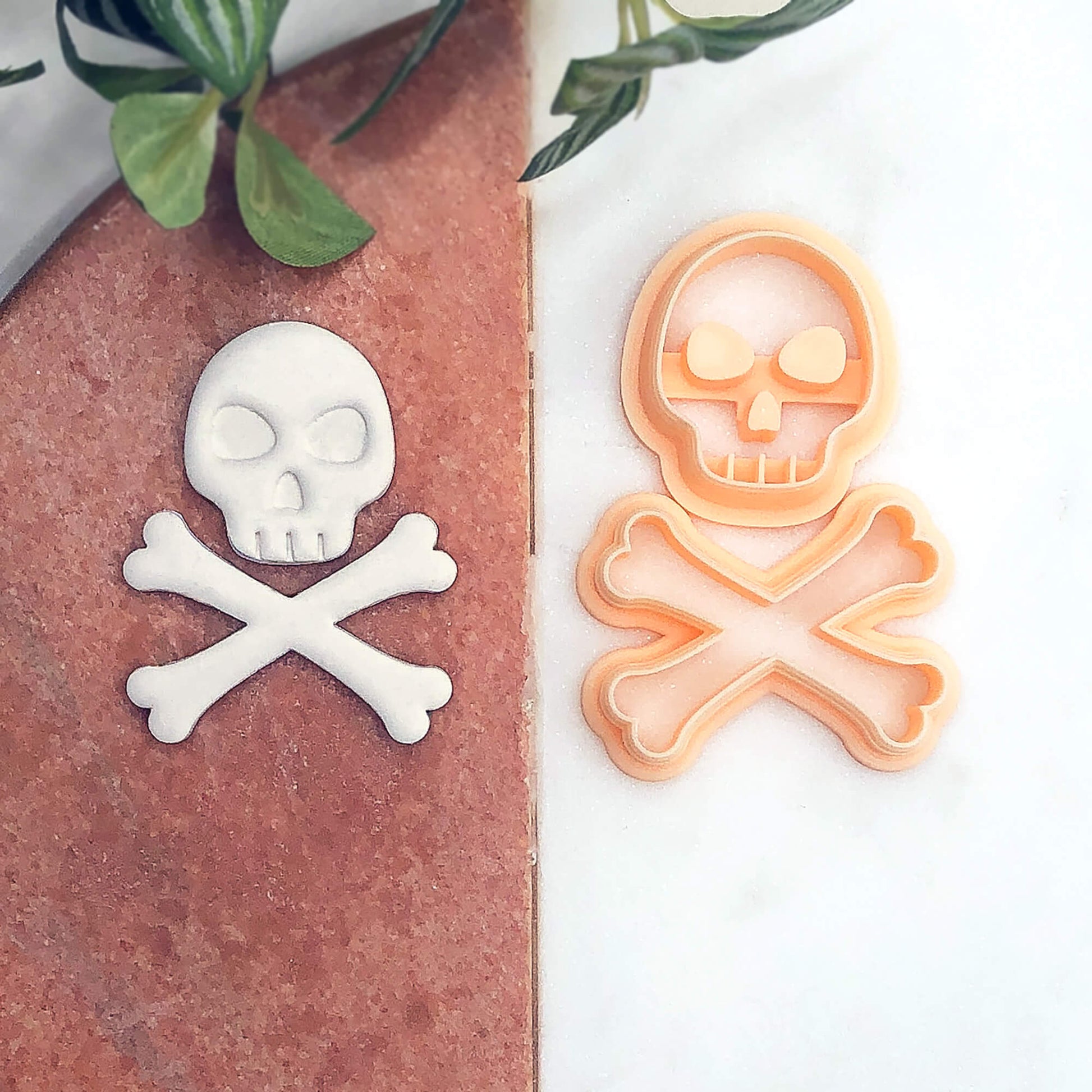 A skull and crossbones cookie cutter set shown on a split background, with a white fondant skull and crossbones piece on a terracotta side and the peach-colored plastic cutter on a white marble side.